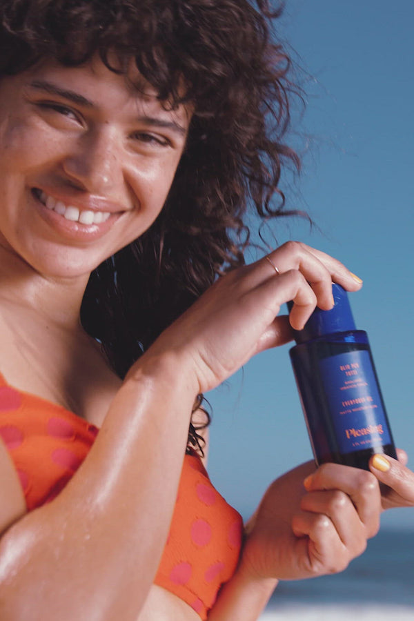 A woman in a bikini holds a bottle of the Everybody Oil Matte Moisture Glow and poses on the beach.